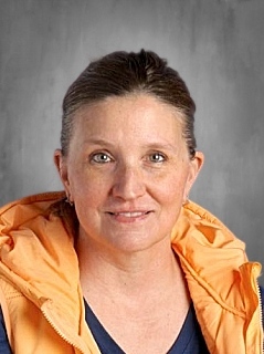 A woman with shoulder-length hair, wearing an orange vest over a blue shirt, smiles against a gray background.