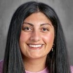 Smiling woman with long black hair, wearing a pink shirt, against a blurred gray background.