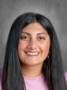 Smiling woman with long black hair, wearing a pink shirt, against a blurred gray background.