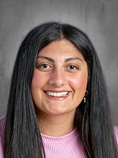 Smiling woman with long black hair, wearing a pink shirt, against a blurred gray background.