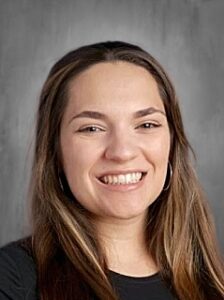 A smiling woman with long, straight hair and hoop earrings poses against a gray background.
