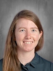 Smiling woman with long brown hair, wearing a dark shirt, against a grey background.