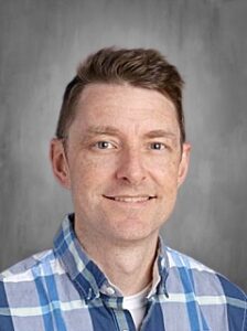 A smiling man with short hair wearing a blue and white plaid shirt against a gray background.