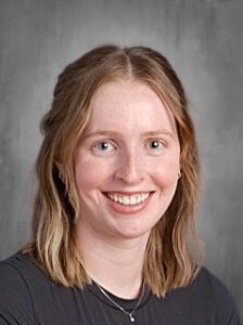 Smiling young woman with medium-length wavy hair, wearing a dark shirt, against a gray background.