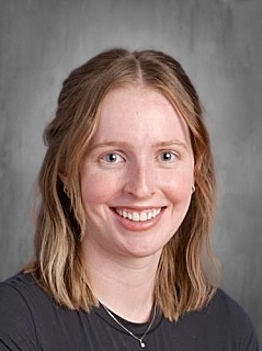 Smiling young woman with medium-length wavy hair, wearing a dark shirt, against a gray background.