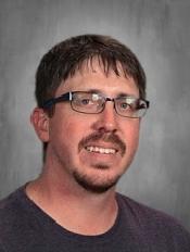 A man with short brown hair and glasses smiles against a gray background. He wears a dark shirt.