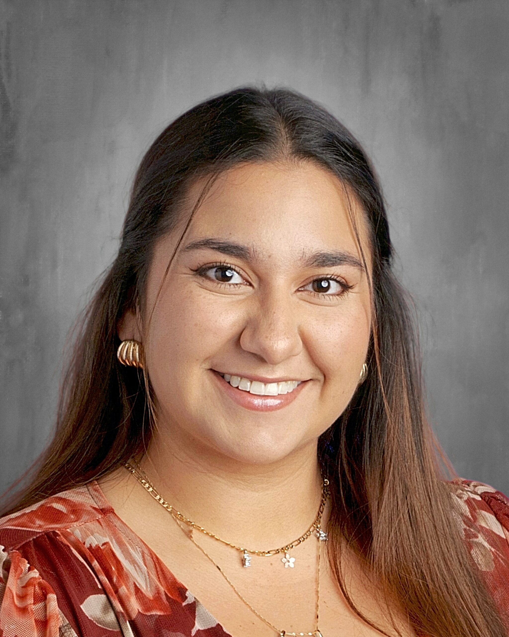 Smiling woman with long dark hair, wearing floral attire and layered necklaces, against a gray background.