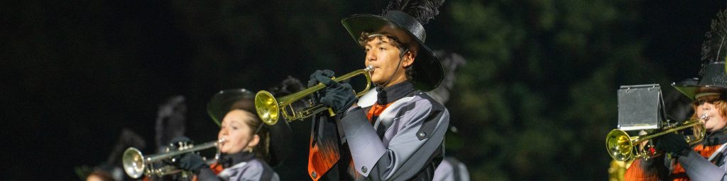 Marching band members in uniform play trumpets outdoors at night.
